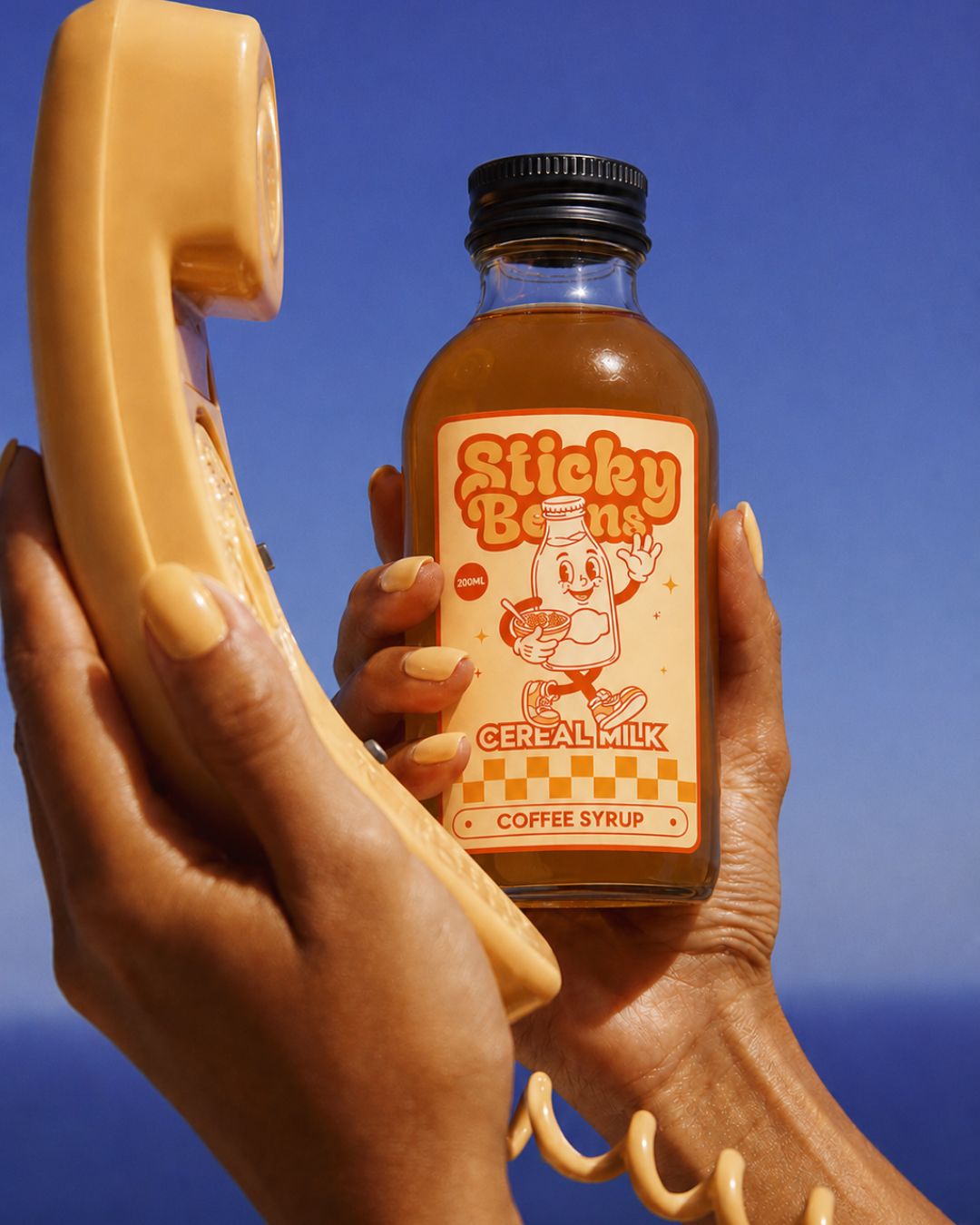 Hands holding a Sticky Beans Cereal Milk coffee syrup bottle next to a vintage phone